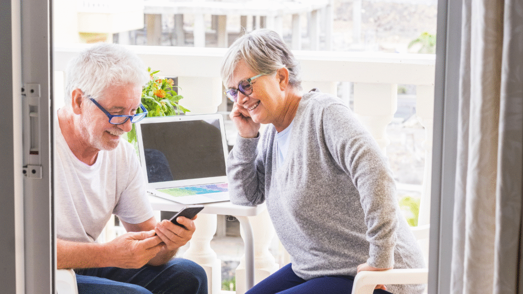 Ein Senior und eine Seniorin sitzen auf einem Balkon und schauen lachend auf ein Smartphone