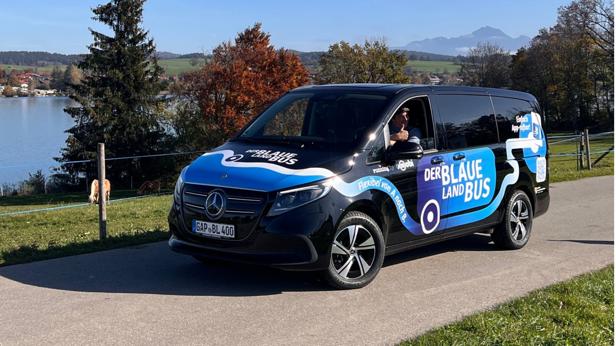 Blaue Land Bus. Ein schwarzer Van steht auf einer kleinen Straße. Im Hintergrund ein See.