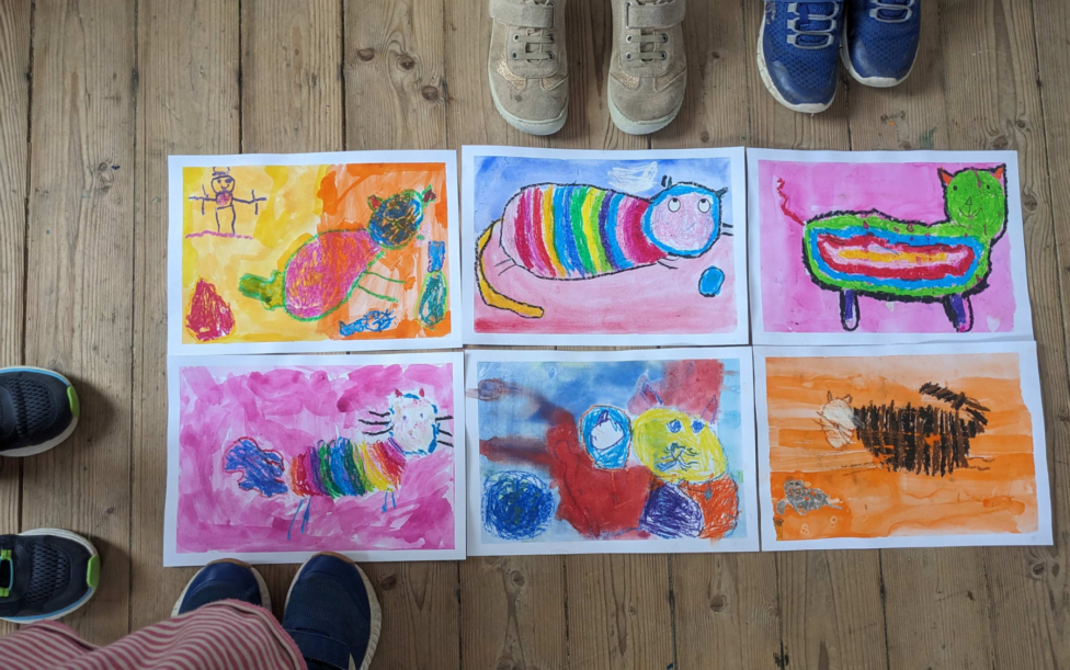 Sechs gemalte Aquarellbilder mit Katzenmotiven liegen auf dem Holzfußboden. Außerdem sind Kinderfüße zu sehen, die drum herum stehen. Ferienprogramm Schlossmuseum Murnau (c) Schlossmuseum Murnau