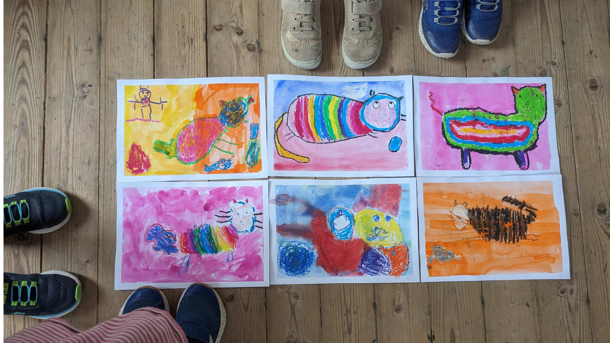 Sechs gemalte Aquarellbilder mit Katzenmotiven liegen auf dem Holzfußboden. Außerdem sind Kinderfüße zu sehen, die drum herum stehen. Ferienprogramm Schlossmuseum Murnau (c) Schlossmuseum Murnau