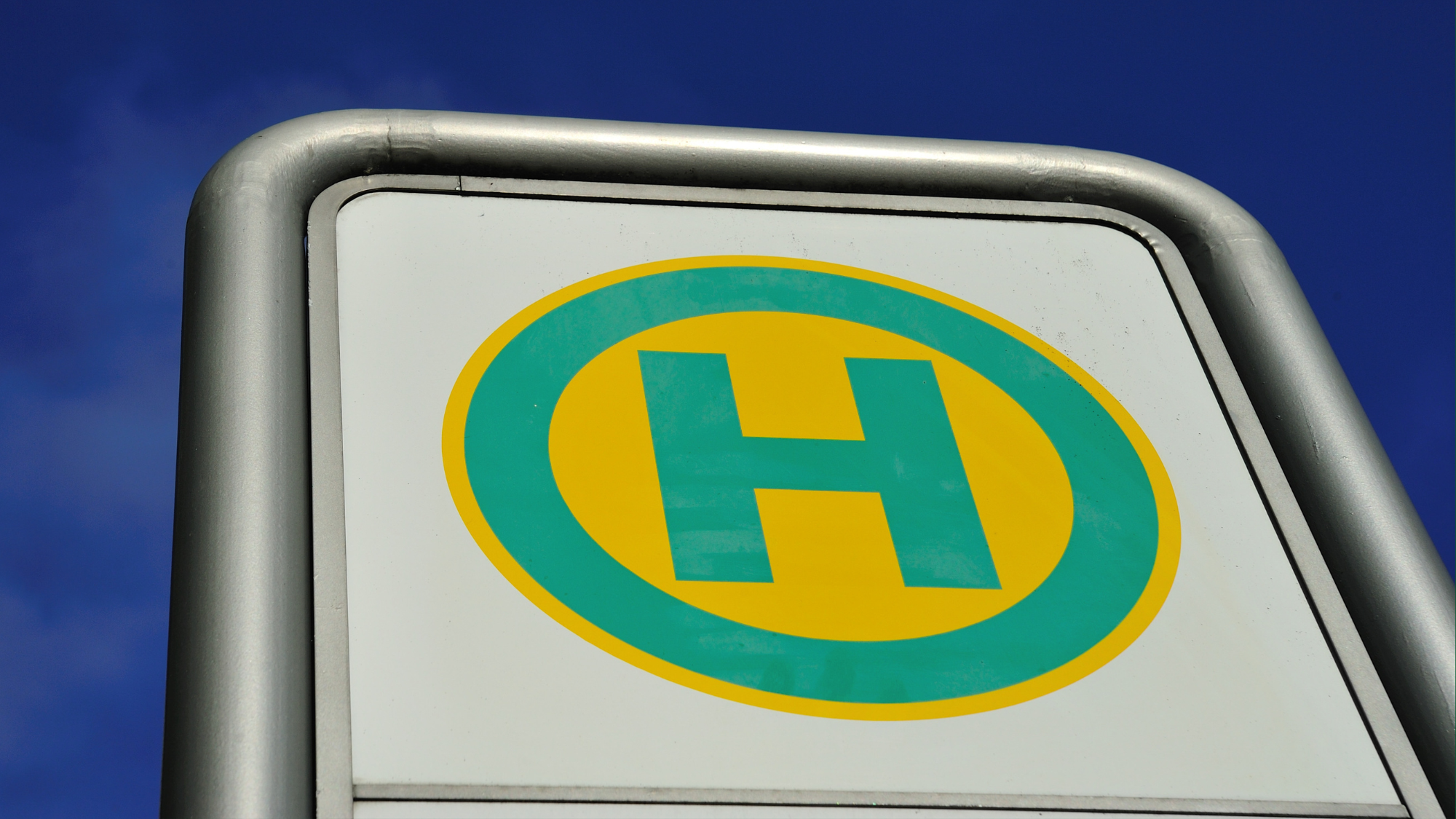 Bushaltestellenschild. Ein grünes H auf gelebn Hintergrund. Hinter dem Schild blauer Himmel.