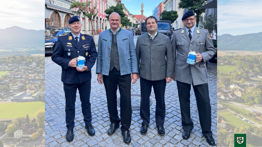 Kriegsgräberfürsorge 2026. Vier Männer, darunter zwei in Militäruniformen und zwei in zivilen Jacken, stehen lächelnd in der Murnauer Fußgängerzone und halten jeweils einen blauen Becher mit weißem Muster.